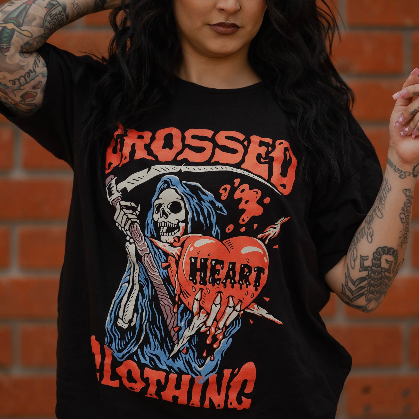 Woman wearing black alternative streetwear t-shirt featuring grim reaper holding heart graphic by Crossed Heart Clothing against brick wall