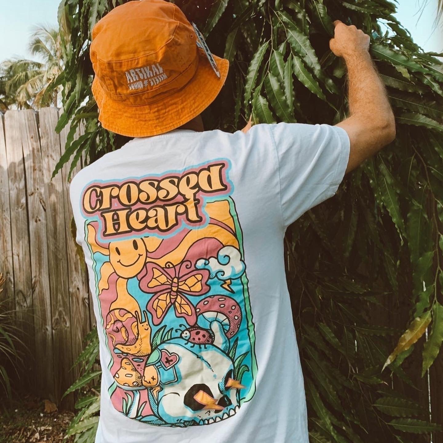 Person wearing white alternative streetwear t-shirt with colorful skull and butterfly back graphic by Crossed Heart Clothing outdoors