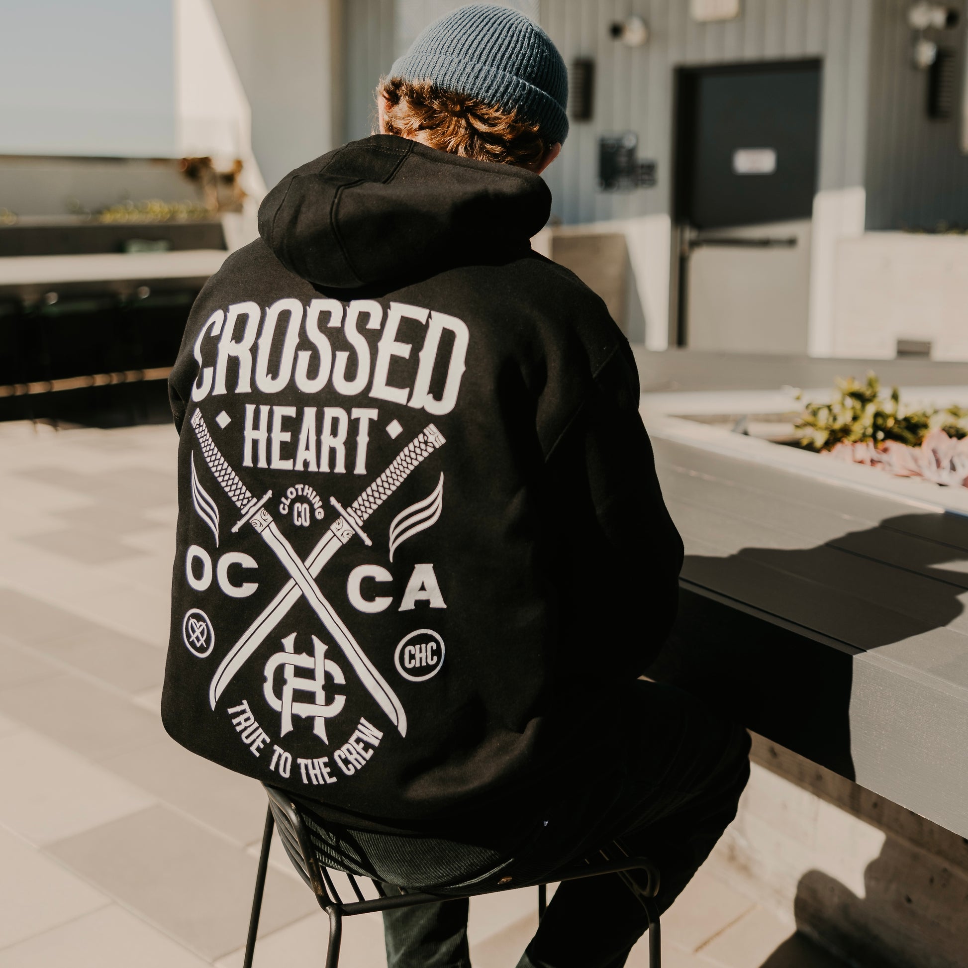 person wearing true to the crew crossed heart black hoodie with oc ca crossed swords graphic tattoo inspired alternative streetwear hoodie outdoors